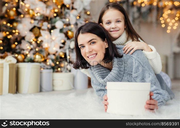 Brunette smiling female adult and her beautiful adorable small kid in white sweater being in living room, hold present, going to congratulate father and husband with coming New Year or Christmas