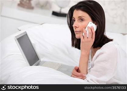 brunette woman at phone and behind a laptop on a bed