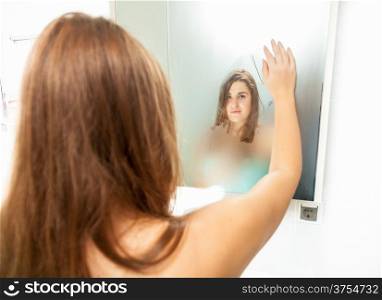 Brunette woman cleaning misted mirror and looking at reflection