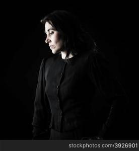 Brunette woman in black standing in front of black backdrop, with a hand on her hip and looking down with a serious, sad expression on her face