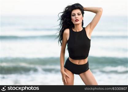 Brunette woman, model of fashion, wearing black top and panties in front of a beach. Young girl with curly hairstyle with eyes closed and moving hair.