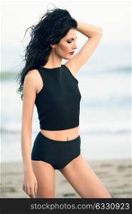 Brunette woman, model of fashion, wearing black top and panties in front of a beach. Young girl with curly hairstyle moving hair.