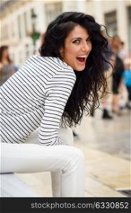 Brunette woman, model of fashion, wearing casual white clothes laughing in the street. Young girl with curly hairstyle sitting in urban background.