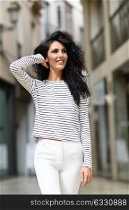 Brunette woman, model of fashion, wearing casual white clothes smiling in the street. Young girl with curly hairstyle standing in urban background.