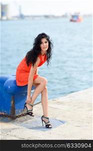 Brunette woman, model of fashion, wearing orange short dress touching her shoe. Young girl with curly hairstyle happy in an urban beach. Female with long legs.