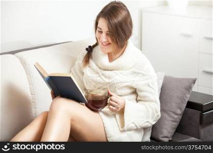 Brunette woman reading funny book on sofa and drinking tea
