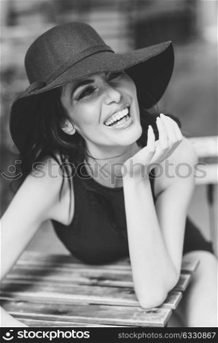 Brunette woman wearing black seductive dress and sun hat laughing in a bar in the street. Young girl with curly hairstyle in urban background