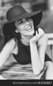 Brunette woman wearing black seductive dress and sun hat laughing in a bar in the street. Young girl with curly hairstyle in urban background