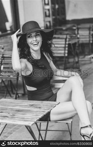 Brunette woman wearing black seductive dress and sun hat sitting in a bar in the street. Young girl with curly hairstyle smiling in urban background