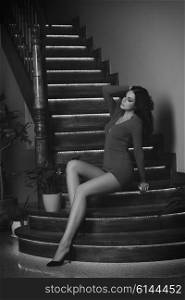 brunette woman with long black hair and sexy red dress in fashion pose sitting on elegant wood stairs . the imagrs is in black and white