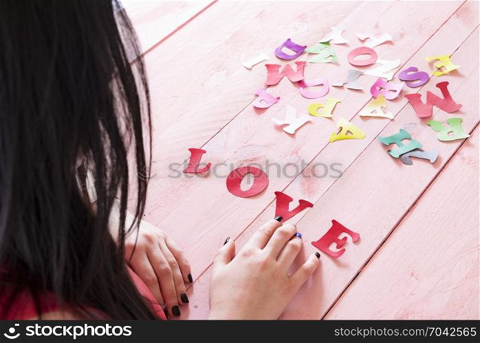 Brunette woman with long hair writing the word love from red paper letters selected from a pile of multicolor letters, on a pink wooden table.