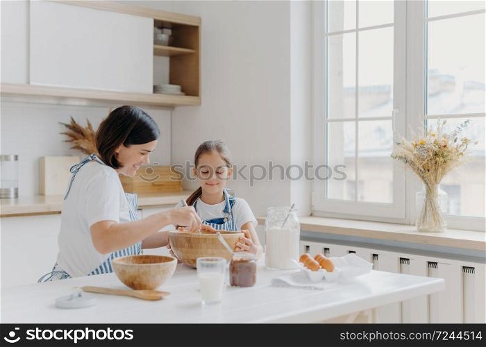Brunette woman with smile shows little daughter how to cook, gives culinary lesson, bought different products in shop for cooking, wear aprons, smile pleasantly, window with vase, kitchen interior