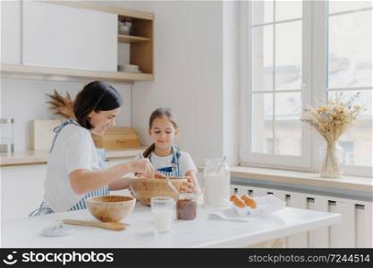 Brunette woman with smile shows little daughter how to cook, gives culinary lesson, bought different products in shop for cooking, wear aprons, smile pleasantly, window with vase, kitchen interior
