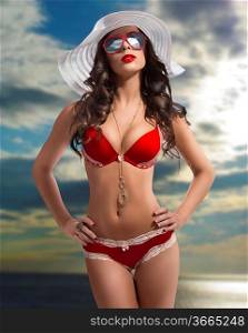 brunette woman with very sexy bikini, big sunglasses and summer white hat with cloudy sky background