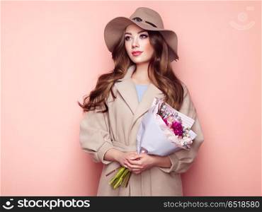 Brunette young woman in stylish greatcoat. Brunette Young Woman in Stylish Greatcoat and Hat. Girl in Beige Coat with Bouquet of Flowers. Model with Long Curly Hair. Spring Lookbook