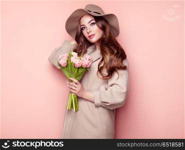 Brunette young woman in stylish greatcoat. Brunette Young Woman in Stylish Greatcoat and Hat. Girl in Beige Coat with Bouquet of Flowers. Model with Long Curly Hair. Spring Lookbook
