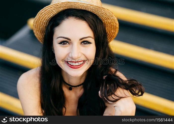 Brunette young woman with attractive appearance, having dark eyes, red painted lips, smiling pleasantly into camera, wearing straw hat sitting at stairs. Beautiful woman showing her positive emotions