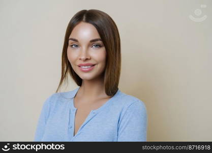 Brunette young woman with healthy beauty skin, smiles gently, shows white perfect teeth, wears minimal makeup, dressed in casual jumper, looks at camera and smiles, isolated on beige background.
