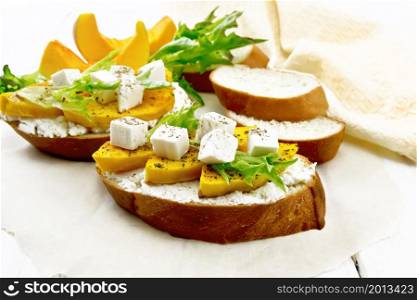 Bruschetta with baked pumpkin, salted feta cheese, ricotta, arugula and spices on parchment, napkin and vegetable slices on the background of light wooden board