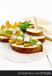 Bruschetta with baked pumpkin, salted feta cheese, ricotta, arugula and spices on parchment, towel and vegetable slices on wooden board background