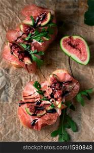 Bruschetta with prosciutto ham, ricotta cheese, arugula and fresh figs on a dark paper background. Appetizing appetizer, starters concept. Flat lay