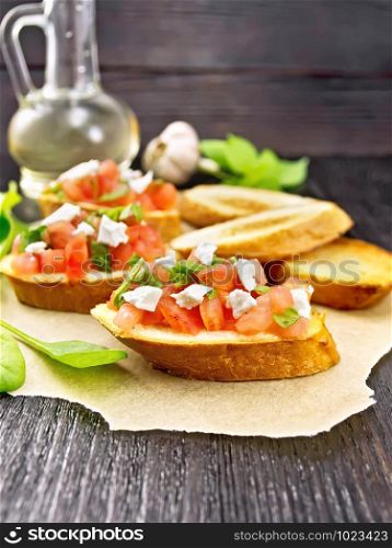 Bruschetta with tomato, spinach and soft cheese on parchment, garlic, vegetable oil in a decanter and basil on wooden board background