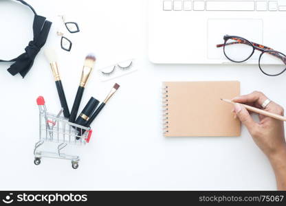 Brushes set in shopping cart, woman accessories and laptop on white background with woman hand holding pencil on blank notebook