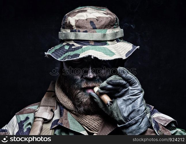 Brutal and serious commando soldier, army special forces veteran, in camouflage battle uniform, boonie hat, black paint on bearded face, combat knife in shoulder holder, smoking cigar, studio portrait. Commando soldier in boonie hat smoking cigar