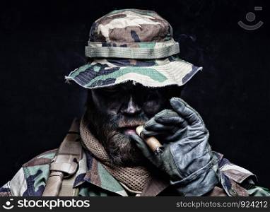 Brutal and serious commando soldier, army special forces veteran, in camouflage battle uniform, boonie hat, black paint on bearded face, combat knife in shoulder holder, smoking cigar, studio portrait. Commando soldier in boonie hat smoking cigar