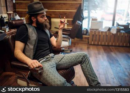Brutal bearded barber in hat with straight razor in hand sitting in a leather chair, barbershop inerior on background