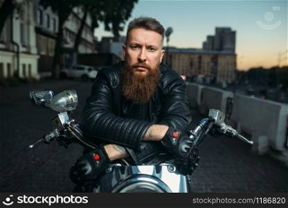 Brutal bearded biker poses on chopper in city, front view. Vintage bike, rider on motorcycle. Brutal bearded biker poses on chopper, front view