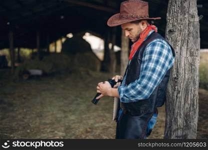 Brutal cowboy checks his revolver before gunfight, texas ranch on background, western. Vintage male person with gun on farm, wild west lifestyle