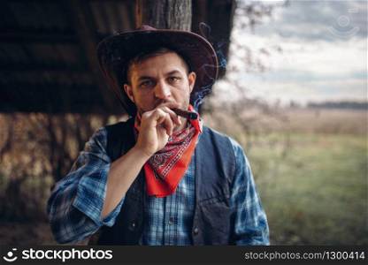 Brutal cowboy smokes a cigar, texas ranch on background, western. Vintage male person with gun on farm, wild west culture. Brutal cowboy smokes a cigar, wild west culture