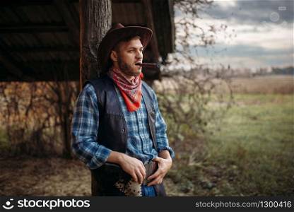 Brutal cowboy smokes a cigar, texas ranch on background, western. Vintage male person with gun on farm, wild west. Brutal cowboy smokes a cigar, wild west culture