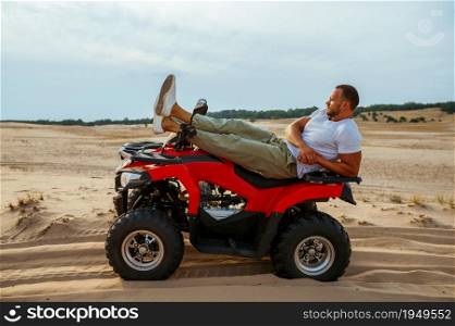 Brutal man lying on atv in desert, side view. Male person on quad bike, sandy race, dune safari in hot sunny day, 4x4 extreme adventure, quad-biking. Brutal man lying on atv in desert, side view
