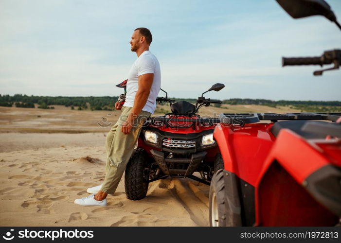Brutal man poses near atv in desert sands. Male person on quad bike, sandy race on quadbike, dune safari in hot sunny day, 4x4 extreme adventure, quad-biking. Brutal man poses near atv in desert, quadbike