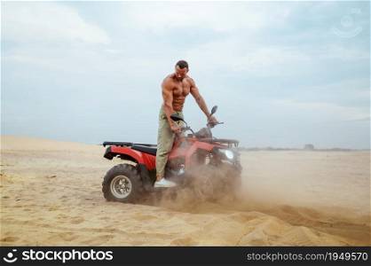 Brutal man with naked body rides on atv in desert sands, quadbike racer. Male person on quad bike, sandy race on quadbike, dune safari in hot sunny day, 4x4 extreme adventure, quad-biking. Brutal man with naked body rides on atv in desert