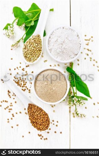 Buckwheat flour from brown and green cereals in two bowls, spoons with different buckwheat groats, flowers and leaves on the background of light wooden board from above