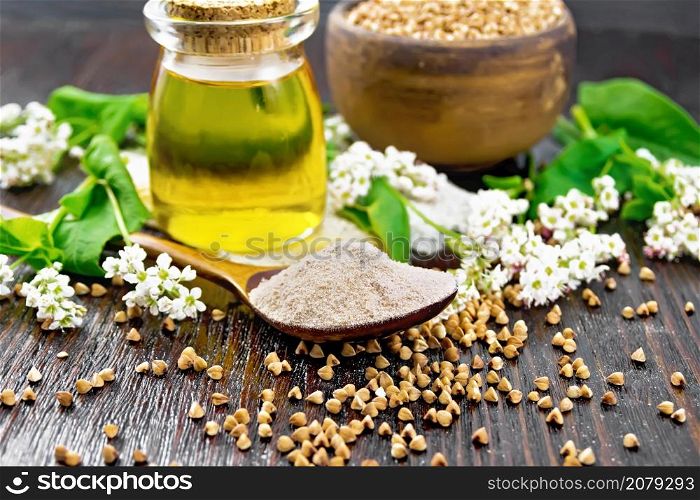 Buckwheat flour from brown cereals in a spoon, oil in a glass jar on sacking, flowers and buckwheat leaves on wooden board background