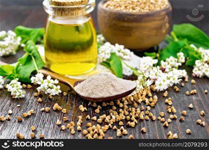 Buckwheat flour from brown cereals in a spoon, oil in a glass jar on sacking, flowers and buckwheat leaves on wooden board background