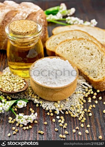 Buckwheat flour from green cereals in a bowl on sacking, buckwheat groats in a spoon and on the table, oil in a glass jar, bread, fresh flowers and leaves on dark wooden board background