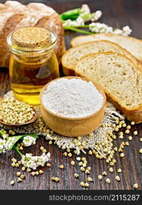 Buckwheat flour from green cereals in a bowl on sacking, buckwheat groats in a spoon and on the table, oil in a glass jar, bread, fresh flowers and leaves on dark wooden board background