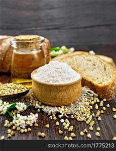 Buckwheat flour from green cereals in bowl on sacking, buckwheat groats in a spoon and on the table, oil in a glass jar, bread, flowers and leaves on wooden board background
