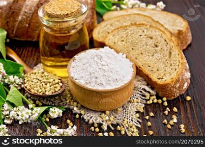 Buckwheat flour from green cereals in bowl on sacking, groats in a spoon and on the table, oil in a glass jar, bread, fresh flowers and buckwheat leaves on a dark background of wooden board
