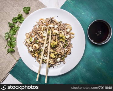 Buckwheat noodles with squid and seafood on a white plate with chopsticks, soy sauce on wooden rustic background top view