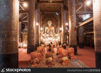 Buddhist monks and novices evening prayer chanting in the ancient temple of Wat Xieng Thong, Luang Prabang, Laos. Soft focus on Buddha statue.
