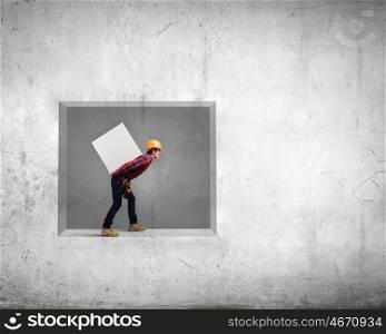 Builder carrying cube. Young builder man in hardhat carrying white blank cube on back