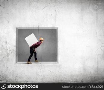 Builder carrying cube. Young builder man in hardhat carrying white blank cube on back