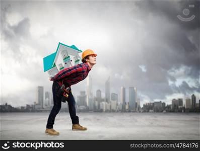 Builder carrying house. Young builder man in hardhat carrying house model on back