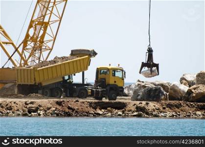Building a dike. Cranes and excavator put stones in the sea. Trucks carrying stones